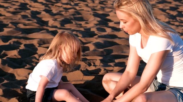 Schöne-blonde-Mutter-und-Tochter-spielen-mit-Sand-sitzen-zusammen-am-Strand.