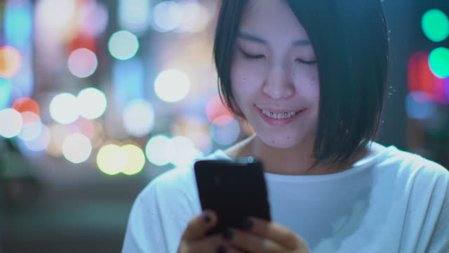 Porträt-der-attraktiven-japanischen-Mädchen-tragen-legere-Kleidung-nutzt-Smartphone.-Im-Hintergrund-Plakate-Großstadt-Werbung-Lichter-leuchten-in-der-Nacht.