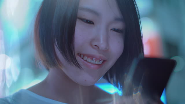 Porträt-von-die-attraktive-Japanerin-mit-Piercing-und-lässige-Kleidung-nutzt-Smartphone.-Im-Hintergrund-Plakate-Großstadt-Werbung-Lichter-leuchten-in-der-Nacht.