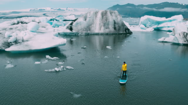 Aerial-shot-of-man-paddling-stand-up-paddle-board-in-glacier-lagoon-with-giant-icebergs-in-Iceland