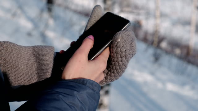 Close-up-woman's-hands-in-mittens-writing-messages-on-cellphone-in-winter-day-in-park.