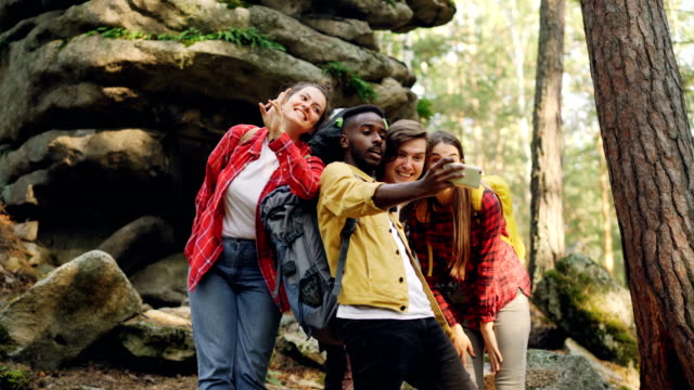 Multiracial-group-of-friends-tourists-are-taking-selfie-in-forest-with-rocks-in-background-using-smartphone,-men-and-women-are-posing-and-showing-hand-gestures.