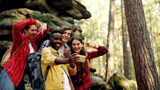 Happy-African-American-man-tourist-is-taking-selfie-with-friends-in-forest-near-huge-rocks-using-smartphone,-people-are-making-funny-faces-and-showing-hand-gestures.