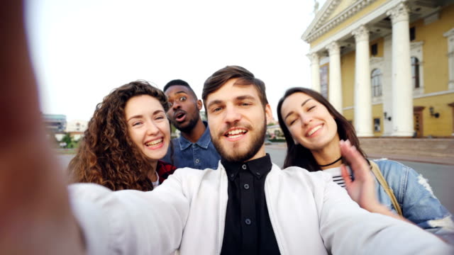 Point-of-view-shot-of-multiracial-group-of-tourists-taking-selfie-in-city-center-holding-camera-and-posing-together-with-hand-gestures-expressing-positive-emotions.