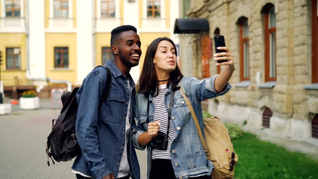 Beautiful-multiracial-couple-is-talking-with-friends-online-making-video-call-using-smartphone-standing-in-the-street-with-backpacks.-Travelling-and-technology-concept.
