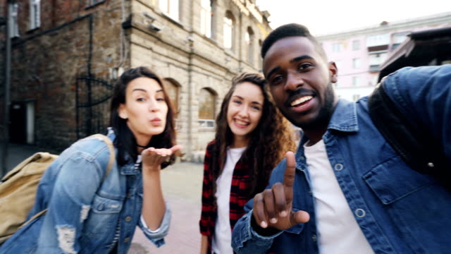 Point-of-view-shot-of-attractive-young-men-and-women-travelers-taking-selfie-in-the-street-posing-and-laughing-holding-camera.-Modern-technology-and-travelling-concept.