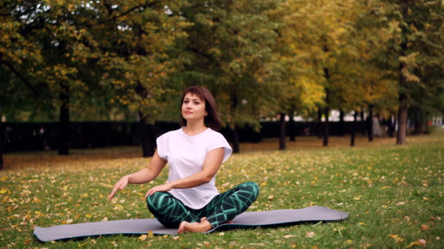 Chica-flexible-yogini-es-practicando-torcedura-simple-pose-luego-reposo-en-posición-de-loto,-sentado-en-la-estera-de-yoga-en-pasto-en-el-parque.-Recreativo-concepto-área-y-la-gente.