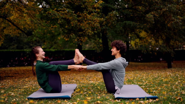 Glückliche-junge-Frauen-tun-Paar-Yoga-mit-Spaß-und-Lachen-auf-Matten-auf-dem-Rasen-im-Park-sitzen.-Schönen-Herbst-Natur-Rasen-und-Bäume-sind-im-Hintergrund.