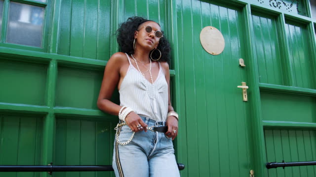 Fashionable-young-black-woman-wearing-sunglasses-leaning-by-green-door-outdoors-looking-at-camera,-low-angle