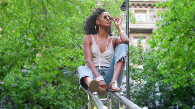 Trendige-junge-schwarze-Frau-trägt-Leibchen-und-Seite-Streifen-Jeans,-sitzt-am-Handlauf-in-grüne-Stadtstraße