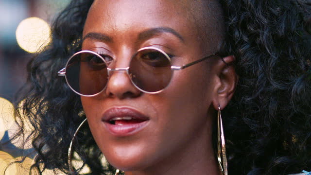 Young-black-woman-wearing-round-sunglasses,-turning-around-and-laughing,-close-up-head-shot,-bokeh-lights-in-background