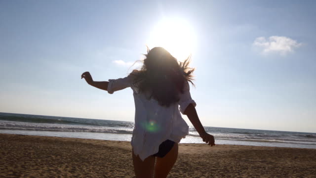 Young-woman--running-on-beach-to-the-ocean-at-sunset-and-raised-hands.-Beautiful-young-girl-going-on-sandy-shore-to-the-sea-and-enjoying-freedom-during-vacation.-Relax-on-summer-holiday.-Rear-back-view