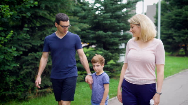 Close-up-of-happy-family-walking-together-on-the-street-laughing.-Father-and-mother-speaking-to-each-other-and-son.-Loving-couple-and-their-child-stepping-on-the-path-with-trees-in-the-background