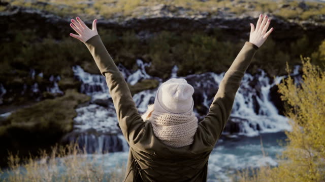 Libertad:-mujer-joven-de-pie-cerca-de-cascadas-de-Islandia-y-levantando-las-manos,-lleva-sombrero-y-esta-feliz