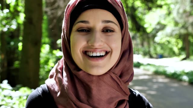 Porträt-von-einem-netten-jungen-Mädchen-in-einem-Hijab,-lächelt,-schaut-in-die-Kamera,-Park-im-Hintergrund.-50-fps