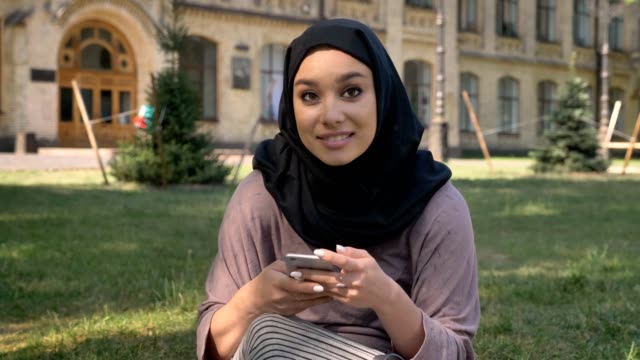 Young-beautiful-muslim-girl-in-hijab-is-watching-at-smartphone-in-daytime-in-summer,-watching-at-camera,-smiling,-building-on-background,-religiuos-concept,-communication-concept