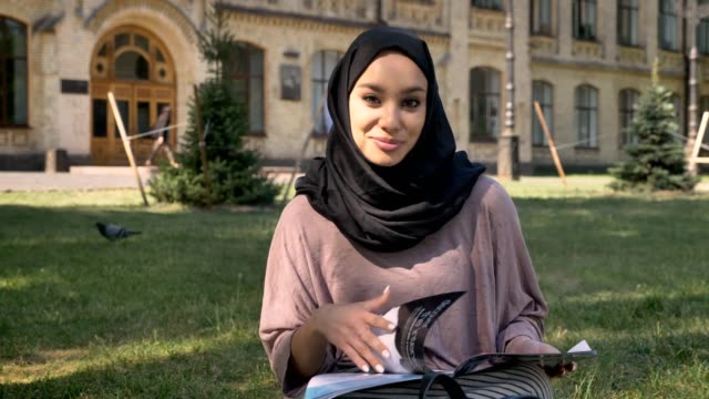 Young-muslim-girl-in-hijab-is-sitting-on-lawn-and-reading-magazine,-watching-at-camera,-smiling,-builging-on-background,-religious-concept,-relax-concept