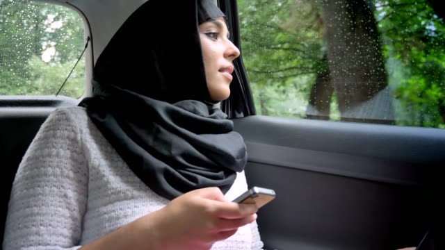 Junge-muslimische-Frau-Hijab-ist-im-verregneten-Fenster-im-Auto,-gerade-halten,-Smartphone,-Transportkonzept,-Kommunikationskonzept,-Wetter-Konzept