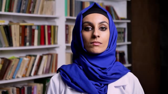 Junge-schöne-Muslima-Hijab-stehen-in-Bibliothek-und-Blick-in-die-Kamera-mit-ernsten-Ausdruck