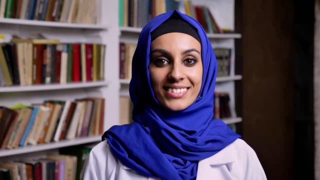Junge-schöne-Muslima-Hijab-in-Bibliothek-steht-und-lächelt-in-die-Kamera-mit-glücklichen-Ausdruck