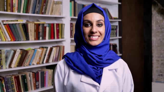 Joven-mujer-musulmana-feliz-en-hijab-en-biblioteca-de-pie-y-mirando-a-cámara-y-sonriendo