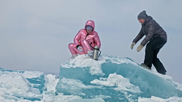 Junges-Paar-hat-Spaß-beim-Winterspaziergang-Hintergrund-von-Eis-der-zugefrorenen-See.-Liebhaber-sind-sitzen-auf-großen-blauen-Eisscholle,-küssen-und-umarmen,-Tee-aus-der-Thermoskanne-trinken.-Liebesgeschichte.