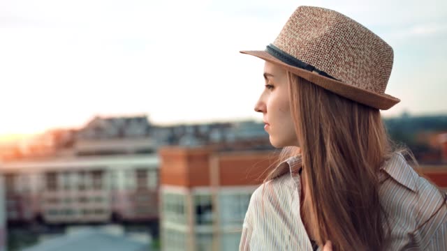 Side-view-melancholic-woman-in-hat-enjoying-city-landscape-at-amazing-sunset-on-roof-or-balcony