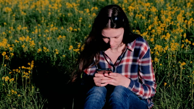 Woman-looking-at-mobile-phone-sitting-in-Park-on-grass-among-yellow-flowers.