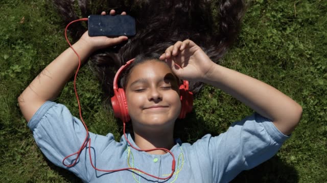 Chica-joven-con-largo-cabello-negro-escuchando-música-en-auriculares-acostado-sobre-su-espalda-en-el-parque-en-tiempo-soleado.-4K
