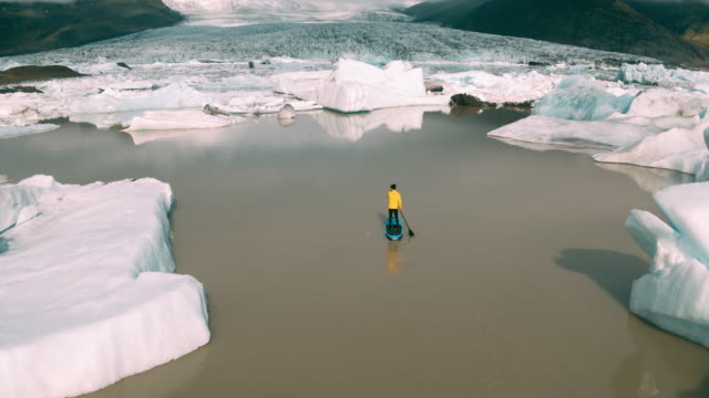 Toma-aérea-del-hombre-remando-de-pie-Junta-de-paleta-en-la-laguna-de-glaciar-con-icebergs-gigantes-en-Islandia