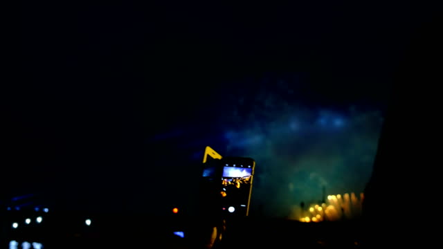 Fireworks-and-crowd-through-the-smartphone-screen.-People-looking-at-the-fireworks-and-pyrotechnic-show-in-the-evening,-bright-flashes-of-light