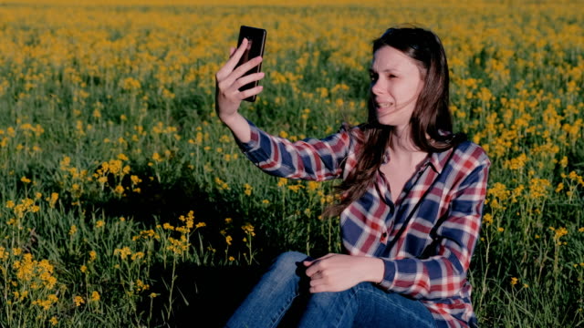 Mujer-hablar-chat-de-video-en-el-teléfono-sentado-en-la-hierba-entre-las-flores-amarillas.