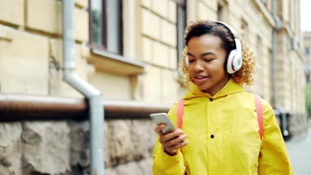 Happy-African-American-girl-is-listening-to-music-through-wireless-headphones-and-using-smartphone-walking-outdoors-enjoying-song-and-walk.-Gadgets-and-millennials-concept.