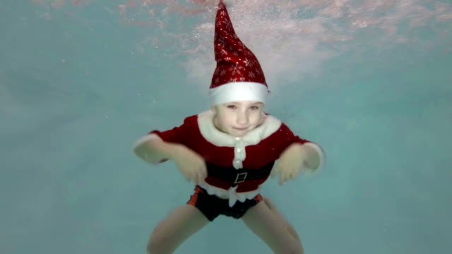 Glücklicher-kleine-Junge-in-einem-roten-Anzug-Weihnachtsmann-schwimmt-unter-Wasser-im-Pool-in-den-Wasserstrahl,-lächelnd-und-Blick-in-die-Kamera.