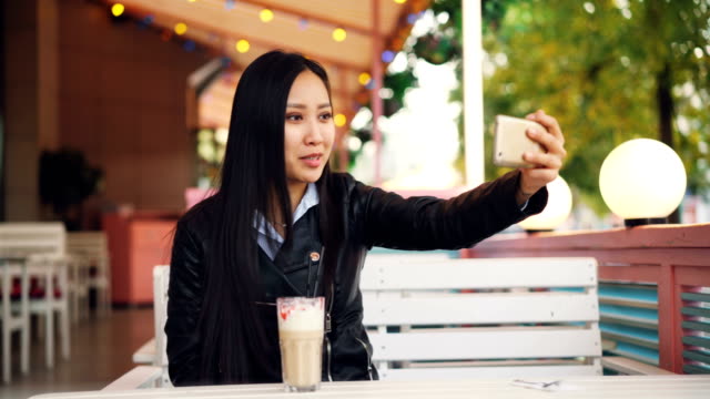 Gerne-asiatische-Dame-spricht-mit-Freunden-online-video-Anruf-mit-Smartphone-in-open-air-Café-allein-sitzen.-Mädchen-spricht,-Haare-zu-berühren-und-lächelnd.