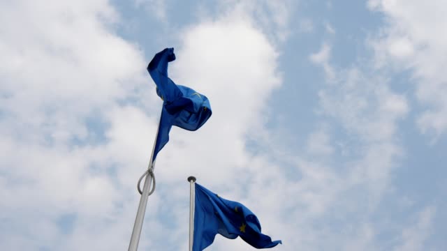Herrliche-Aussicht-auf-zwei-EU-Flaggen,-die-an-einem-sonnigen-Frühlingstag-vor-dem-EU-Parlament-in-Brüssel-wehen.-Der-Himmel-ist-blau-und-bewölkt.
