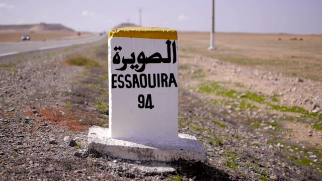 Nahaufnahme-der-Entfernung-Schild-Straße-zu-essaouira-in-Französisch-und-Arabisch-geschrieben-mit-Kutschen-im-Hintergrund.-Marokko