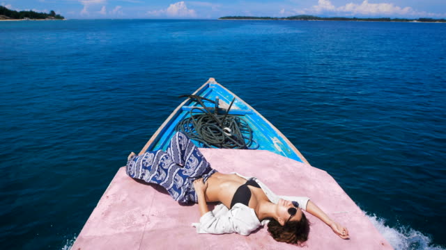 Tanned-young-girl-in-sunglasses-with-a-thrown-back-arm-behind-her-head-is-in-the-stern-of-the-boat,-which-quickly-goes-on-the-dark-blue-ocean-to-a-small-island