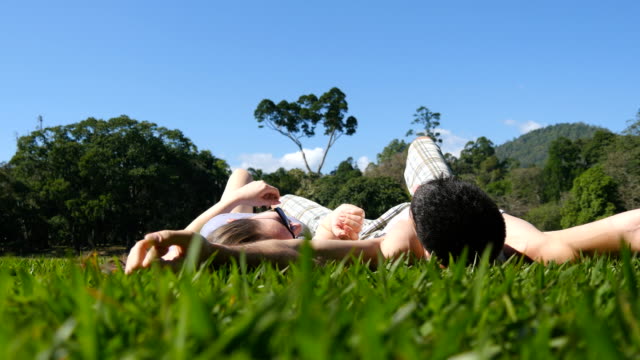 Junges-Paar-am-grünen-Rasen-im-Park-liegen-und-entspannen.-Mann-und-Frau-sitzen-auf-der-Wiese-am-Natur-und-küssen.-Mädchen-und-Jungen-mit-Blick-auf-die-Landschaft-und-Urlaub-genießen.-Geringe-Blickwinkel-hinten-zurück