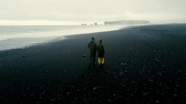 Back-aerial-view-of-the-young-couple-walking-on-the-black-volcanic-beach-in-Iceland.-Romantic-date-near-the-sea