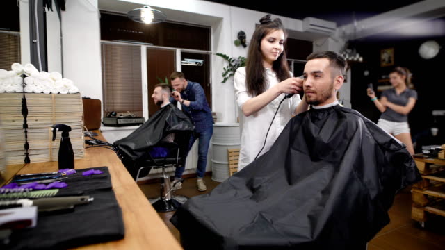 Two-friends-visiting-modern-barbershop.-Handsome-hipsters-sitting-against-mirror-and-working-with-hairstylists.-Smiling-man-and-woman-using-professional-equipment-for-making-stylish-haircuts