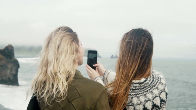 Zwei-glückliche-Frauen-fotografieren-Selfie-auf-Smartphone,-in-der-Nähe-von-Mädchen,-die-Zeit-am-Strand-verbringen-den-Troll-Zehen-in-Island