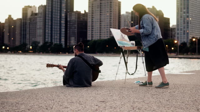 Joven-pareja-creativa-de-artistas-callejeros-haciendo-afición-en-orilla-de-Michigan,-Chicago,-Estados-Unidos.-Sorteos-de-mujer,-hombre-juegos-de-guitarra