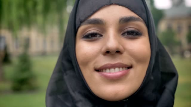 Cara-de-joven-hermosa-chica-musulmana-en-hijab-es-de-pie-y-sonriente-durante-el-día,-mal-tiempo,-mirando-a-cámara,-concepto-de-epoca,-fondo-borroso