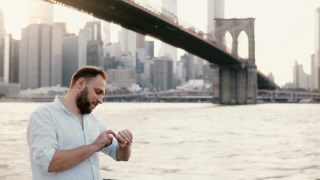 Successful-young-European-businessman-standing-near-river-under-Brooklyn-Bridge,-using-his-smart-watch,-walking-away-4K