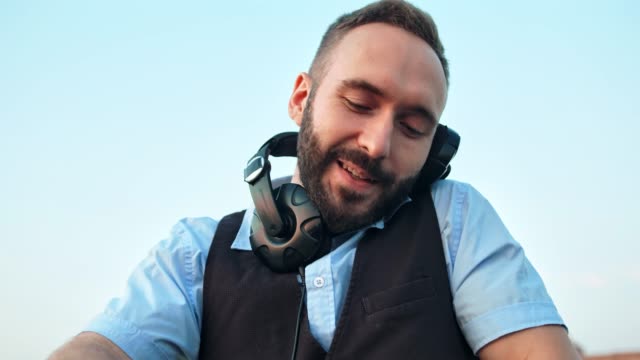 Low-angle-portrait-enthusiastic-happy-male-DJ-in-headphones-at-outdoor-disco-or-rooftop-party