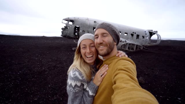 Junges-paar-stehend-durch-Flugzeug-Wrack-am-schwarzen-Sandstrand-unter-Selfie-Porträt-Sehenswürdigkeit-zu-besuchen-in-Island-und-stellen-mit-dem-Wrack---4K