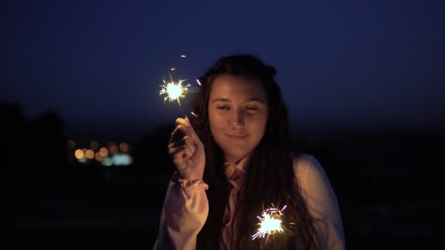 A-young-girl-with-long-dark-hair-stands-against-the-background-of-the-city-at-night-and-holds-fireworks-in-a-good-mood.-slow-motion.-4K