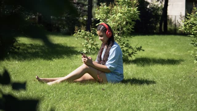 Chica-escuchando-música-en-auriculares-sentado-sobre-la-hierba-en-el-parque.-4K
