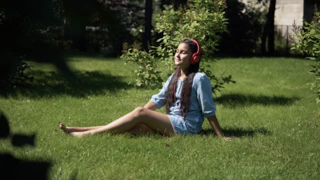 Young-girl-listening-to-music-on-headphones-sitting-on-grass-in-park-in-sunny-weather.-4K
